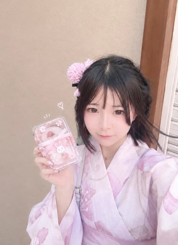 さな(JK上がりたて)🎀(18)が桜色の着物を着て、かわいいお菓子を持ちながら微笑んでいる。